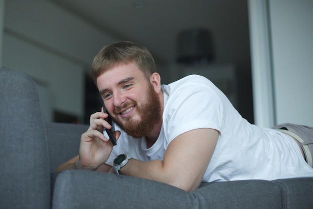 Szczęśliwy młody człowiek rozmawia na czacie przez telefon komórkowy z uśmiechem, baw się dobrze, siedząc na wygodnej kanapie w domu