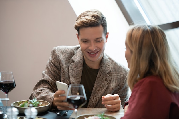 Zdjęcie szczęśliwy młody człowiek w eleganckim ubraniu, patrząc na ekran smartfona, oglądając ciekawe wideo i pokazując go swojej dziewczynie