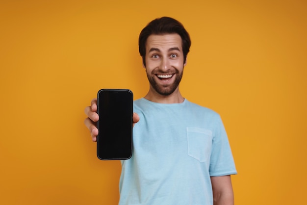 Szczęśliwy młody człowiek w swobodnym ubraniu pokazujący swój inteligentny telefon i uśmiechnięty