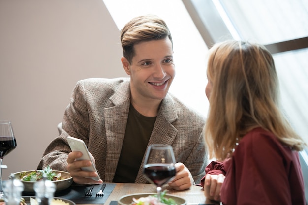 Szczęśliwy młody człowiek ze smartfonem patrzący na swoją dziewczynę z uśmiechem podczas rozmowy z nią przy serwowanym stole w luksusowej restauracji