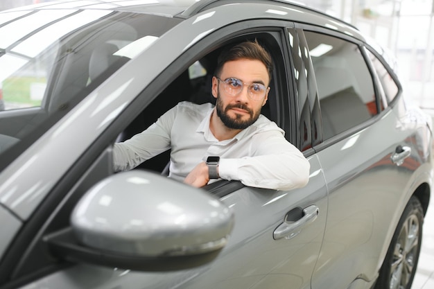 Szczęśliwy młody facet sprawdza nowy luksusowy samochód kupujący samochód w centrum dealerskim Portret wesołego tysiącletniego mężczyzny rasy kaukaskiej badającego auto w salonie