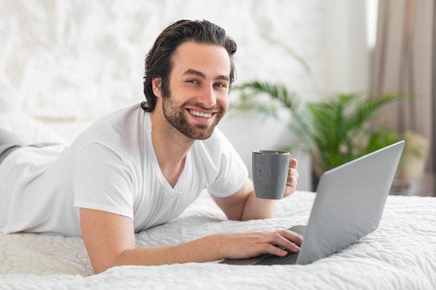 Szczęśliwy młody facet za pomocą laptopa w łóżku pije kawę
