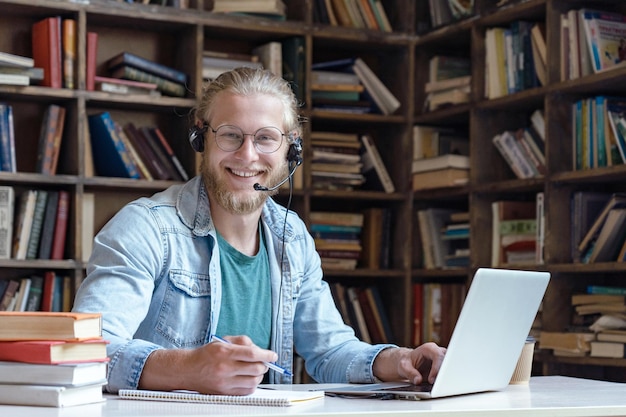 Szczęśliwy młody mężczyzna, student, nosi zestaw słuchawkowy, korzystając z laptopa w portrecie biblioteki