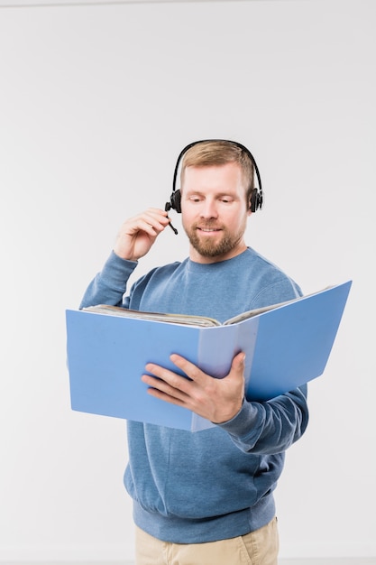 Szczęśliwy młody operator w zestawie słuchawkowym patrząc na jeden z dokumentów w folderze podczas konsultacji z klientami online w izolacji