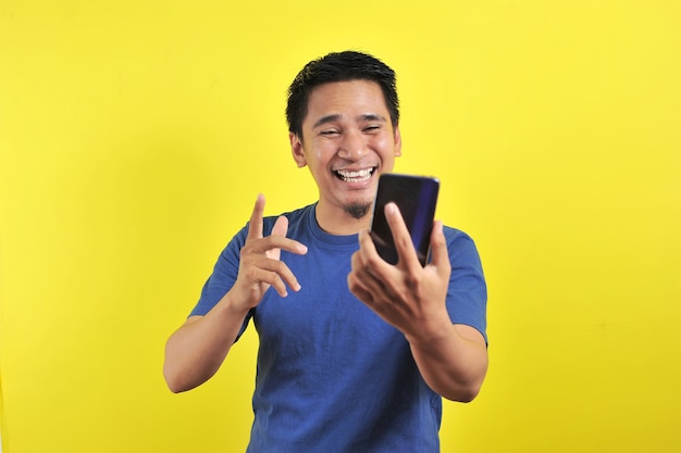 Szczęśliwy młody przystojny Azjata uśmiechający się za pomocą smartfona na żółtym tle