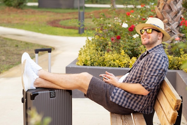 Szczęśliwy młody przystojny mężczyzna rasy kaukaskiej w okularach przeciwsłonecznych z walizką przybywających do hoteli siedzących na ławce w pobliżu hoteluPodróżujący turysta na początku wakacji koncepcji wakacji