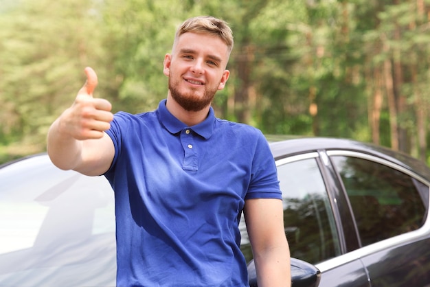 szczęśliwy młody przystojny mężczyzna stojący w pobliżu swojego samochodu uśmiechnięty pokaż kciuk w górę kupując samochód właściciel samochodu
