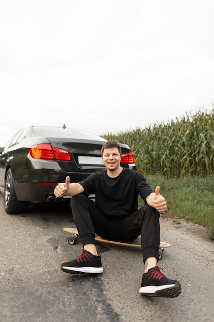 Szczęśliwy Młody Przystojny Mężczyzna W Czarnych Ubraniach Siedzi I Odpoczywa Na Longboardzie W Pobliżu Samochodu I Wskazuje Palcem W Górę Podróżowanie Samochodem I Aktywne Wakacje