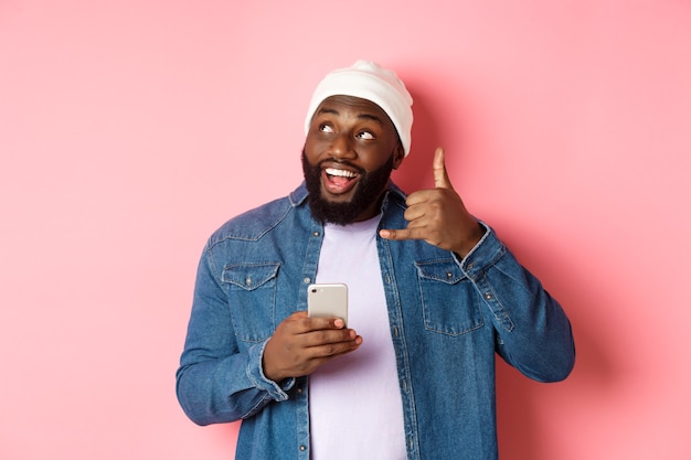 Szczęśliwy Murzyn Pokazując Znak Zadzwoń Do Mnie, Wykonując Gest Telefonu I Uśmiechając Się, Trzymając Smartfon, Stojąc W Czapce I Dżinsowej Koszuli Na Różowym Tle.