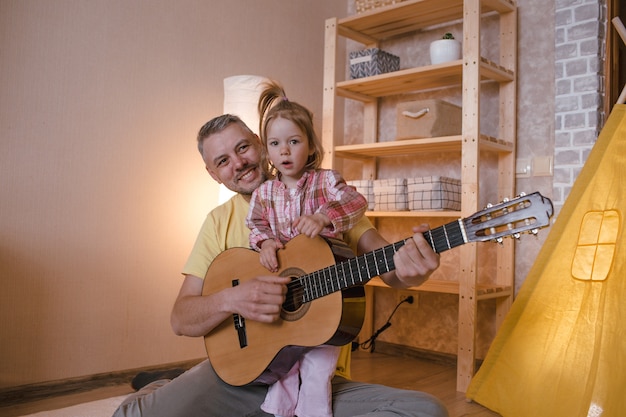 Szczęśliwy ojciec z córeczką uczy się gry na gitarze siedząc w domu na podłodze obok żółtego tipi. szczęśliwy ojciec