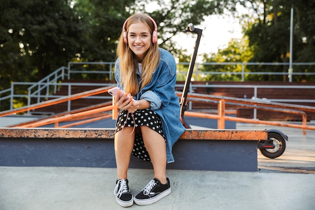 szczęśliwy optymistyczny pozytywny młoda nastolatka poza w parku słuchania muzyki ze słuchawkami trzymając telefon komórkowy.