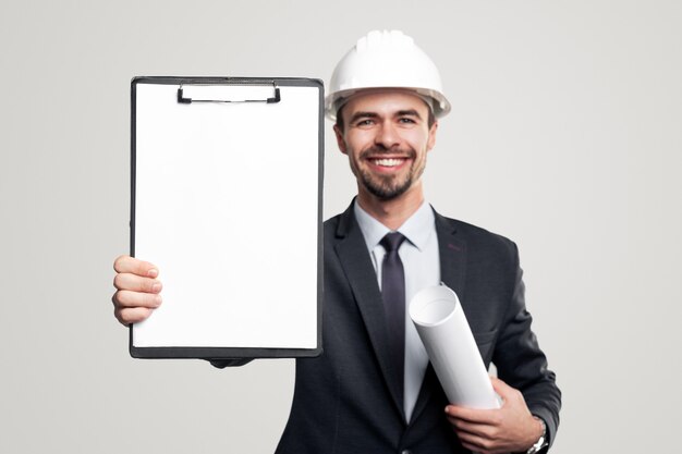 Szczęśliwy, pewny siebie męski inżynier w formalnym garniturze i kasku z zwiniętym planem w ręku, demonstrujący schowek z pustym białym arkuszem papieru