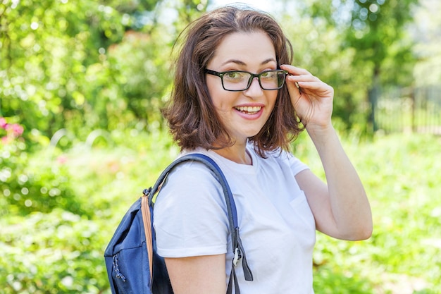 Szczęśliwy Piękny Pozytywny Student Dziewczyna W Okularach Z Plecakiem Uśmiechający Się Na Tle Zielonego Parku. Kobieta Odpoczywająca W Kampusie Podczas Przerwy Na Lunch. Koncepcja Edukacji I Wypoczynku.