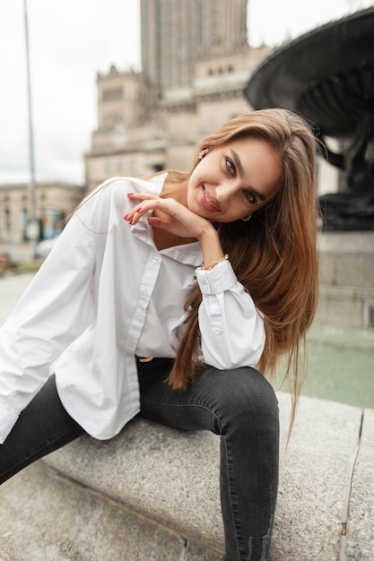 Szczęśliwy piękny stylowy model dziewczyny z uśmiechem w modnych, swobodnych ubraniach z białą koszulą i dżinsami siedzi w pobliżu fontanny w mieście