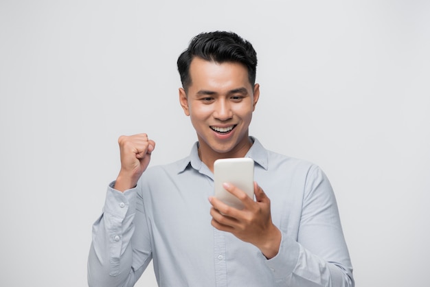 Szczęśliwy podekscytowany Azjata patrzący na swój smartfon i podnoszący rękę, by świętować sukces