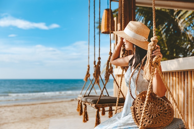 Szczęśliwy podróżnik azjatycka kobieta z kapeluszem i sukienką relaksuje się na huśtawce w kawiarni na plaży, Koh Chang, Tajlandia