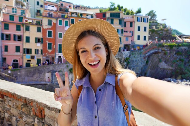 Szczęśliwy podróżnik dziewczyna biorąc selfie zdjęcie robi znak zwycięstwa w słoneczny dzień w wiosce Riomaggiore, Cinque Terre, Włochy