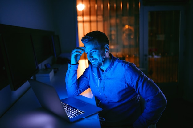 Szczęśliwy pracownik whit toothy uśmiech i słuchawki w ucho używa laptop dla rozmowy wideo podczas gdy siedzący w biurze póżno przy nocą.