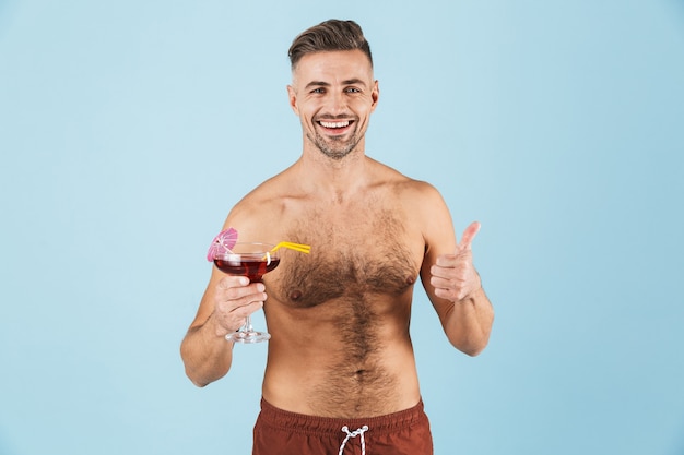 Szczęśliwy przystojny młody przystojny mężczyzna na sobie szorty plażowe, stojąc na niebiesko, pijąc koktajl