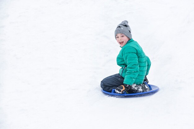 Szczęśliwy roześmiany chłopak ześlizguje się ze wzgórza na śnieżnym spodku. Koncepcja sezonowa. Zimowy dzień.