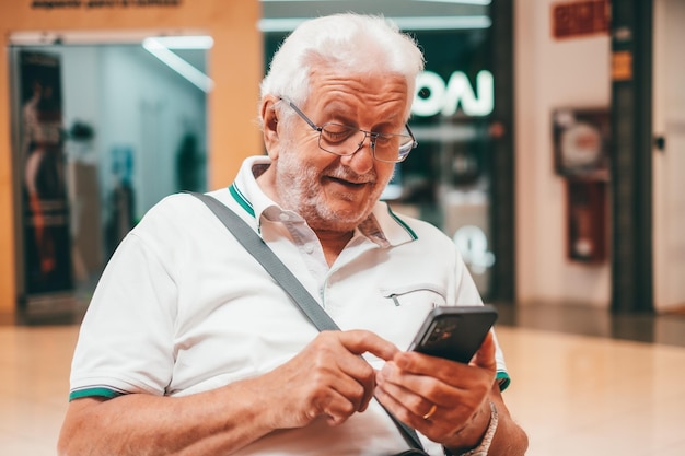 Szczęśliwy starszy mężczyzna siedzi w centrum handlowym za pomocą dziadka smartfona czytającego sms z dobrą wiadomością