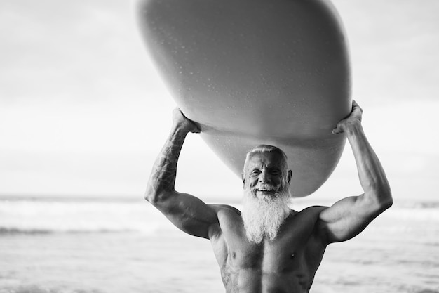 Szczęśliwy starszy surfer trzymający deskę surfingową na plaży o zachodzie słońca Skup się na twarzy Edycja czarno-biała
