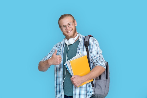 Szczęśliwy student płci męskiej trzyma plecak z kciukami do góry odizolowanymi na niebieskim tle