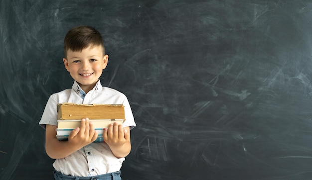 Szczęśliwy student stojący przy tablicy kredowej w klasie z podręcznikami lub książkami w ręku Śmiejący się uczeń szkoły podstawowej Rok akademicki Powrót do szkoły Baner Miejsce na tekst