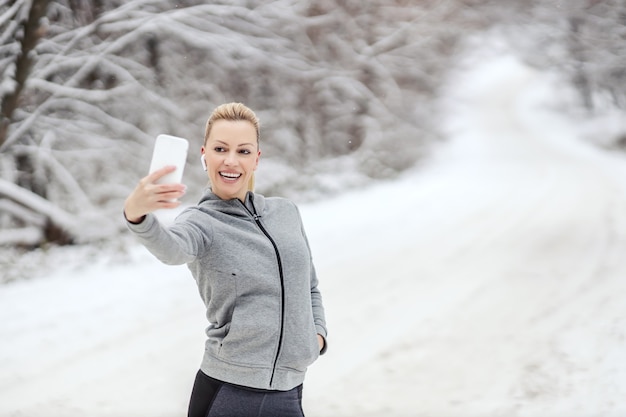 Szczęśliwy szczupły sportsmenka stojący w przyrodzie w mroźny zimowy dzień i przy selfie. Technologia, fitness zimowy