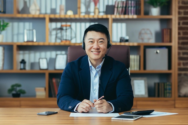 Szczęśliwy szef azjatyckiego biznesmena patrzy w kamerę uśmiechając się i rozmawiając z kolegą podczas konferencji online używa zestawu słuchawkowego mężczyzna w garniturze pracuje w biurze