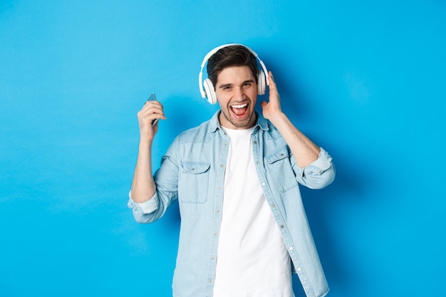 Szczęśliwy uśmiechający się człowiek korzystający ze słuchania muzyki w słuchawkach, trzymając smartfon w uniesionej ręce, stojąc na niebieskiej ścianie