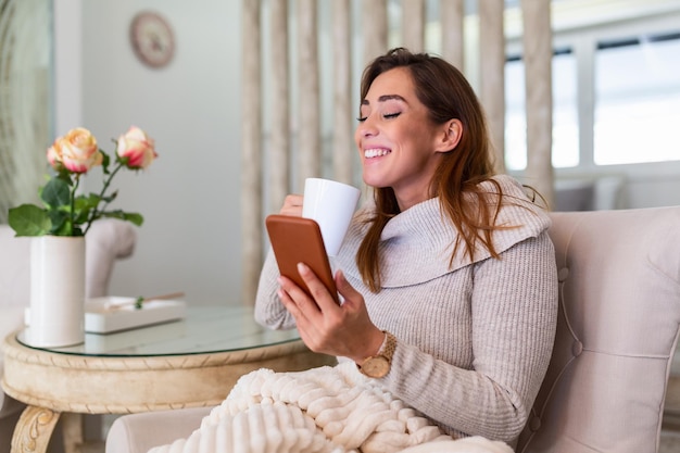 Szczęśliwy uśmiechający się piękna dziewczyna za pomocą urządzenia smartphone w przytulnym domu, atrakcyjna młoda kobieta rozmawia z przyjaciółmi w sieci społecznościowej, siedząc na kanapie w domu i pijąc kawę