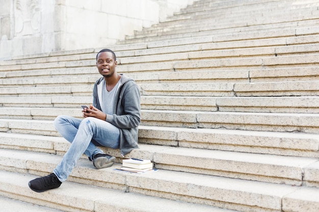 Szczęśliwy uśmiechający się student afro-amerykański siedzi na zewnątrz na schodach uniwersytetu z smartphone, pisanie na klawiaturze, surfowanie po internecie. Koncepcja technologii, edukacji i pracy zdalnej, kopia przestrzeń