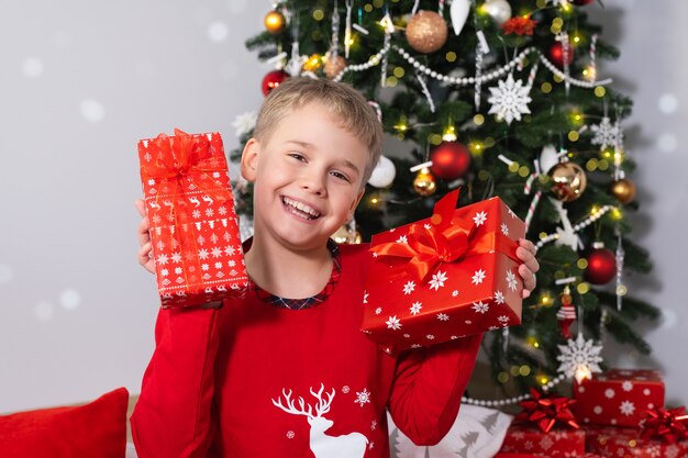 Szczęśliwy uśmiechnięty chłopiec z świątecznymi pudełkami prezentowymi w pobliżu choinki