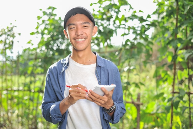 Szczęśliwy uśmiechnięty młody rolnik azjatycki mężczyzna trzyma notatnik na zielonym ogrodzie, na miejscu.