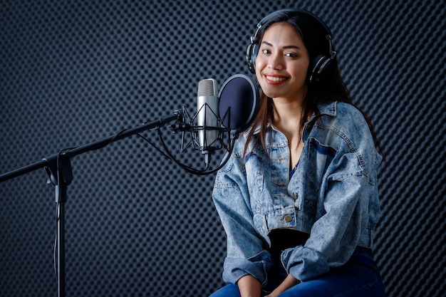 Szczęśliwy wesoły, całkiem uśmiechnięty portret młodej azjatyckiej wokalistki noszącej słuchawki nagrywającej piosenkę przed mikrofonem w profesjonalnym studiu