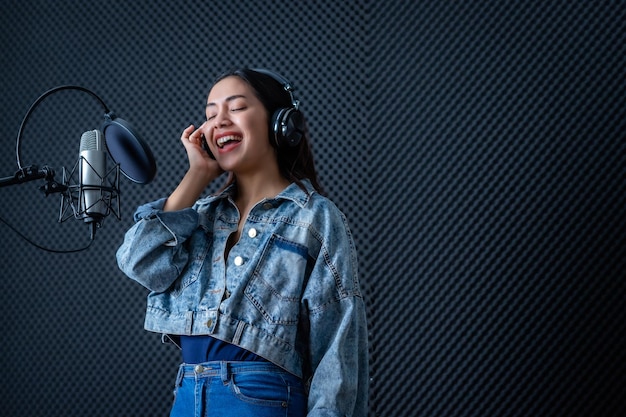 Szczęśliwy, wesoły, całkiem uśmiechnięty portret młodej wokalistki azjatyckiej kobiety w słuchawkach nagrywający piosenkę przed mikrofonem w profesjonalnym studiu