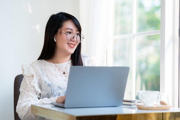 Szczęśliwy z azjatyckich freelancerów Bizneswoman nosi bezprzewodowe słuchawki dorywczo pracując z laptopem z filiżanką kawy Notebook i smartfon w kawiarniKomunikacja w stylu życia biznesowego
