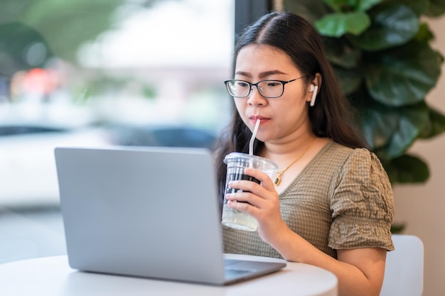 Szczęśliwy z azjatyckich freelancerów Bizneswoman noszący bezprzewodowe słuchawki z ssaniem kawy pracujący z laptopemNotebook i smartfon w kawiarniKomunikacja w stylu życia biznesowego
