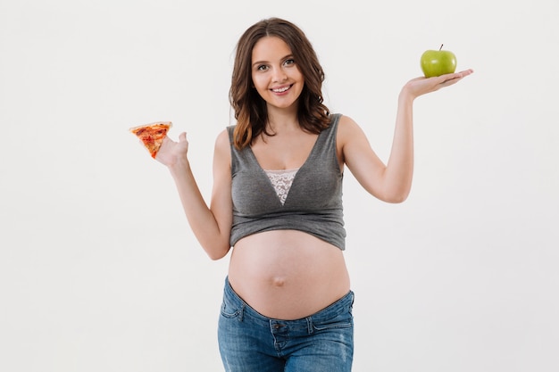 Szczęśliwy zdrowy kobieta w ciąży wybiera między jabłkiem i pizzą.