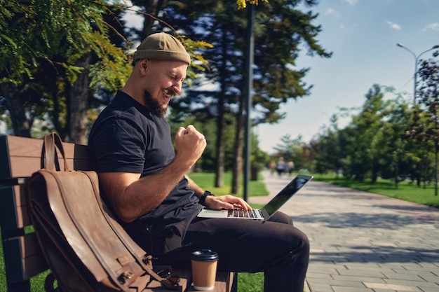 Szczęśliwy Zwycięski Brodaty Mężczyzna Siedzi Na ławce Na świeżym Powietrzu, Patrząc Na Laptopa Gestykulującego Tak, Wygraj Zwycięzcę