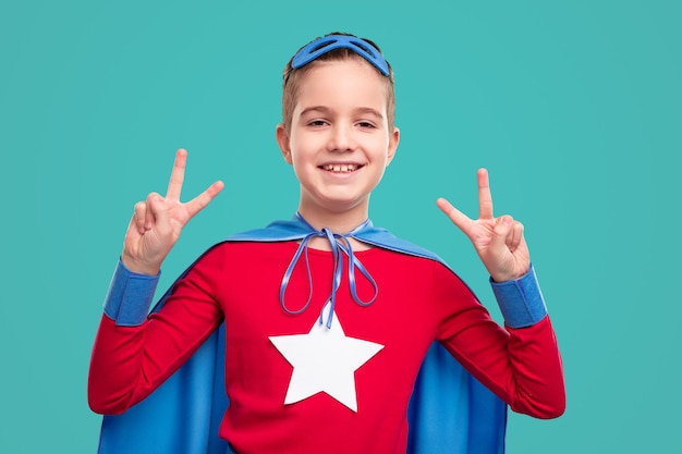 Szczęśliwy zwycięski chłopiec w jasnym kostiumie superbohatera pokazujący znak V jako symbol zwycięstwa i patrzący na kamerę na turkusowym tle
