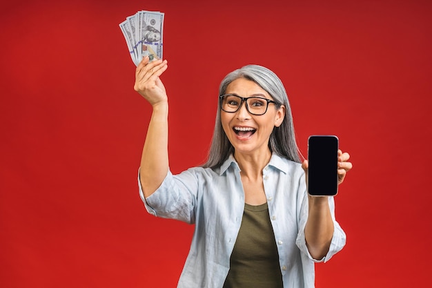 Szczęśliwy zwycięzca Obraz dojrzałej, starzejącej się, szczęśliwej azjatyckiej starszej kobiety stojącej odizolowanej na czerwonym tle ściany trzymającej pieniądze za pomocą telefonu komórkowego Pokazuje ekran telefonu