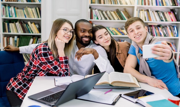 Szczęśliwych Czterech Wieloetnicznych Studentów Wykonujących śmieszne Selfie W Bibliotece Podczas Wspólnego Studiowania. Zabawna Edukacja, Akademia, Szkoła