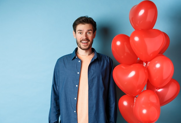 Szczęśliwych walentynek. Podekscytowany uśmiechnięty facet stojący w pobliżu balonów czerwonych serc i patrząc na kamery, niebieskie tło.