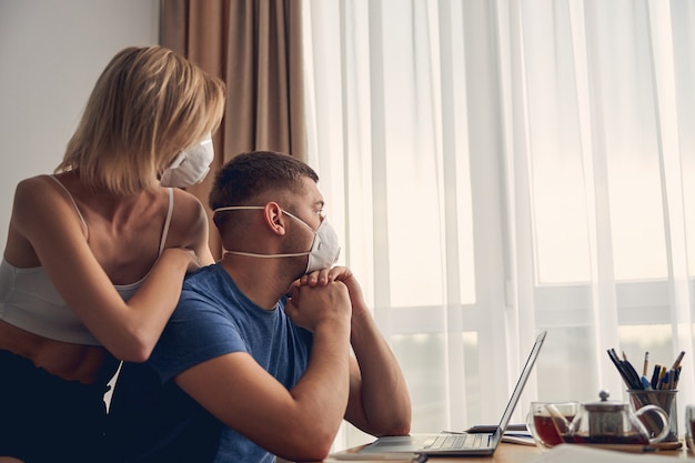 Szczupła blond kobieta opiera się o brunetkę wysportowanego mężczyznę, nosząc maski i patrząc w okno