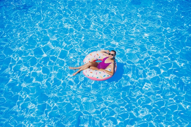 Szczupła kobieta w okularach przeciwsłonecznych w basenie na dmuchanym kółku do pływania w jasnym stroju kąpielowym letnie zdjęcie fotografia pływania letnie zdjęcia kobiety Moda plażowa