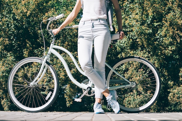 Szczupła młoda kobieta w białej koszulce i dżinsach stoi latem przy swoim stylowym rowerze