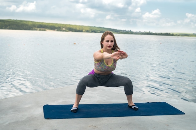 Szczupła, piękna kobieta w strojach sportowych ćwiczy jogi na macie nad jeziorem w słoneczny letni dzień, fitness na świeżym powietrzu. Pojęcie zdrowego stylu życia