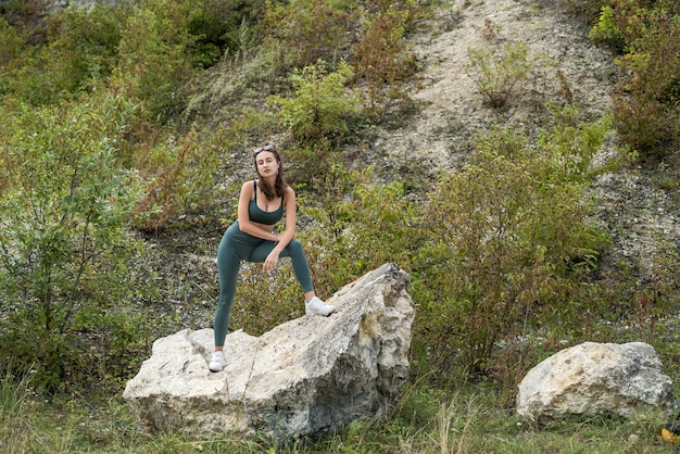 Szczupła seksowna młoda kobieta pozuje w pobliżu dużego kamienia i zielonej wysokiej trawy, idealna letnia przygoda